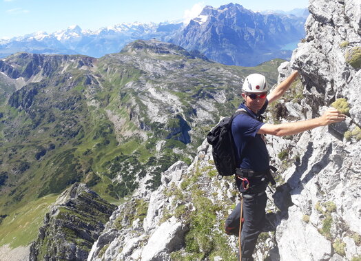Ein Bergsteiger mit weissem Helm befindet sich auf einem Felsband. Im Hintergrund sind die Glarneralpen mit Vrenelisgaertli, Clariden und Toedi zu erkennen.