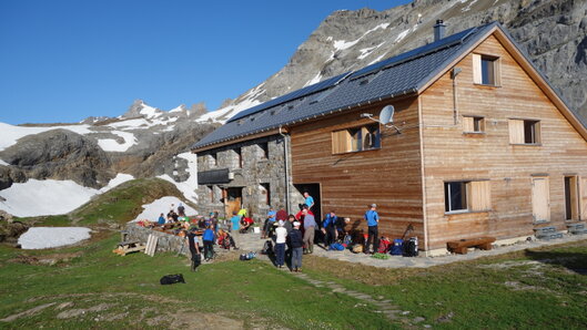 Die Claridenhuette in der Sonne. Vor der Huette befinden sich circa 20 Personen mit Rucksaecken.
