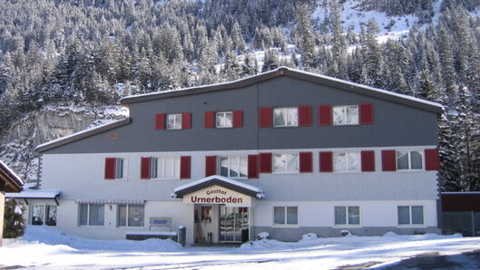 Das Gasthaus Urnerboden mit roten Fensterlaeden, dahinter verschneiter Wald.