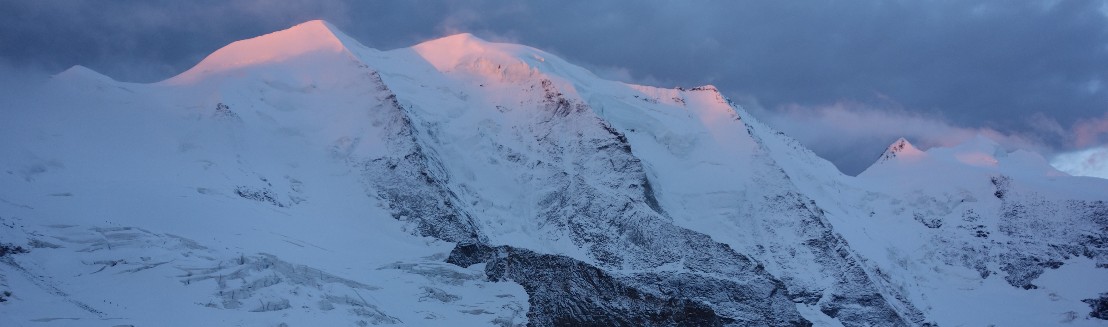 Der Piz Palue mit den drei Gipfeln und den drei Felspfeilern. Die Spitzen der Gipfel werden von der Sonne angeleuchtet und sind orange.