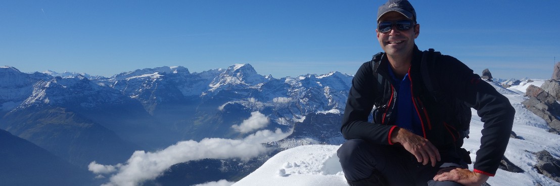 Ein Mann in dunkler Kleidung, posiert knieend und lächelnd auf dem Gipfel des Vrenelisgärtli. Im Hintergrund unter dem blauen Himmel sieht man den Bifertenstock und den Toedi.